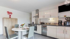 A modern kitchen with white cabinets, a stainless steel range hood, a microwave, and a coffee maker. There is a dining table with two chairs and a vase of flowers.