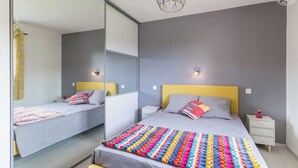 A modern bedroom with a mirrored wall, two beds with colorful bedding, bedside tables, and a unique geometric light fixture.
