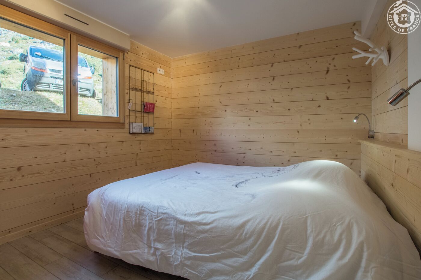Un dormitorio con paredes de madera, una cama, un ventanal y un estante con objetos.