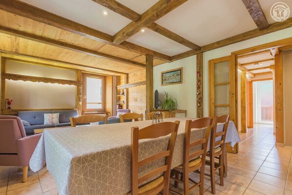 A cozy dining area with a long table and wooden chairs, a sofa, and a television in the background.