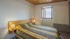 Ein Schlafzimmer mit Holzbalkendecke, einem Bett mit farbenfroher Decke, einer Nachttischlampe und einem Fenster mit Aussicht.