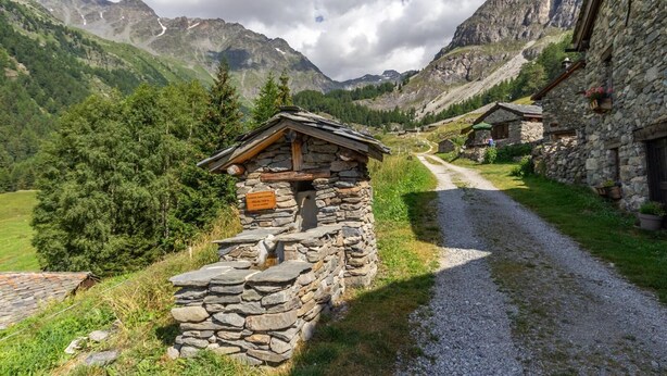 Uma cabana de pedra com telhado de madeira, rodeada por vegetação exuberante e montanhas ao fundo.