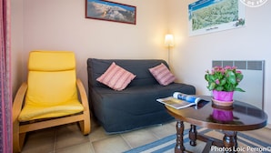 A room with a yellow chair, a dark sofa with patterned pillows, a small round table with a magazine, a potted plant, and two wall-mounted pictures. - BELLE VUE - VEREL PRAGONDRAN (VEREL PRAGONDRAN)