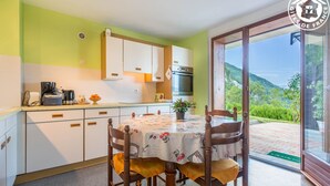 A kitchen with white cabinets, a dining table with chairs, and a view of a green landscape through a glass door. - BELLE VUE - VEREL PRAGONDRAN (VEREL PRAGONDRAN)