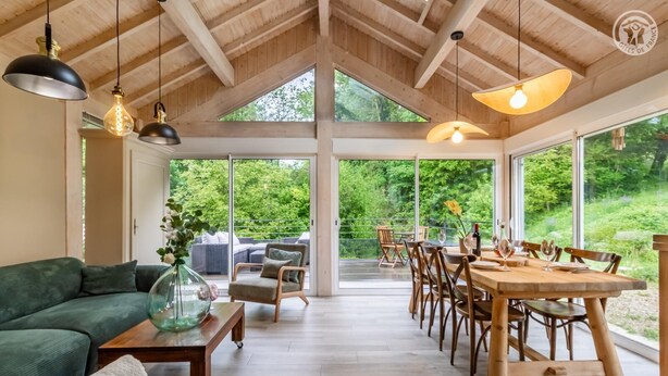A spacious room with a high ceiling, large windows, a dining area with wooden furniture, and a green sofa. - LA BERGERIE - MEYRIEUX TROUET (MEYRIEUX TROUET)