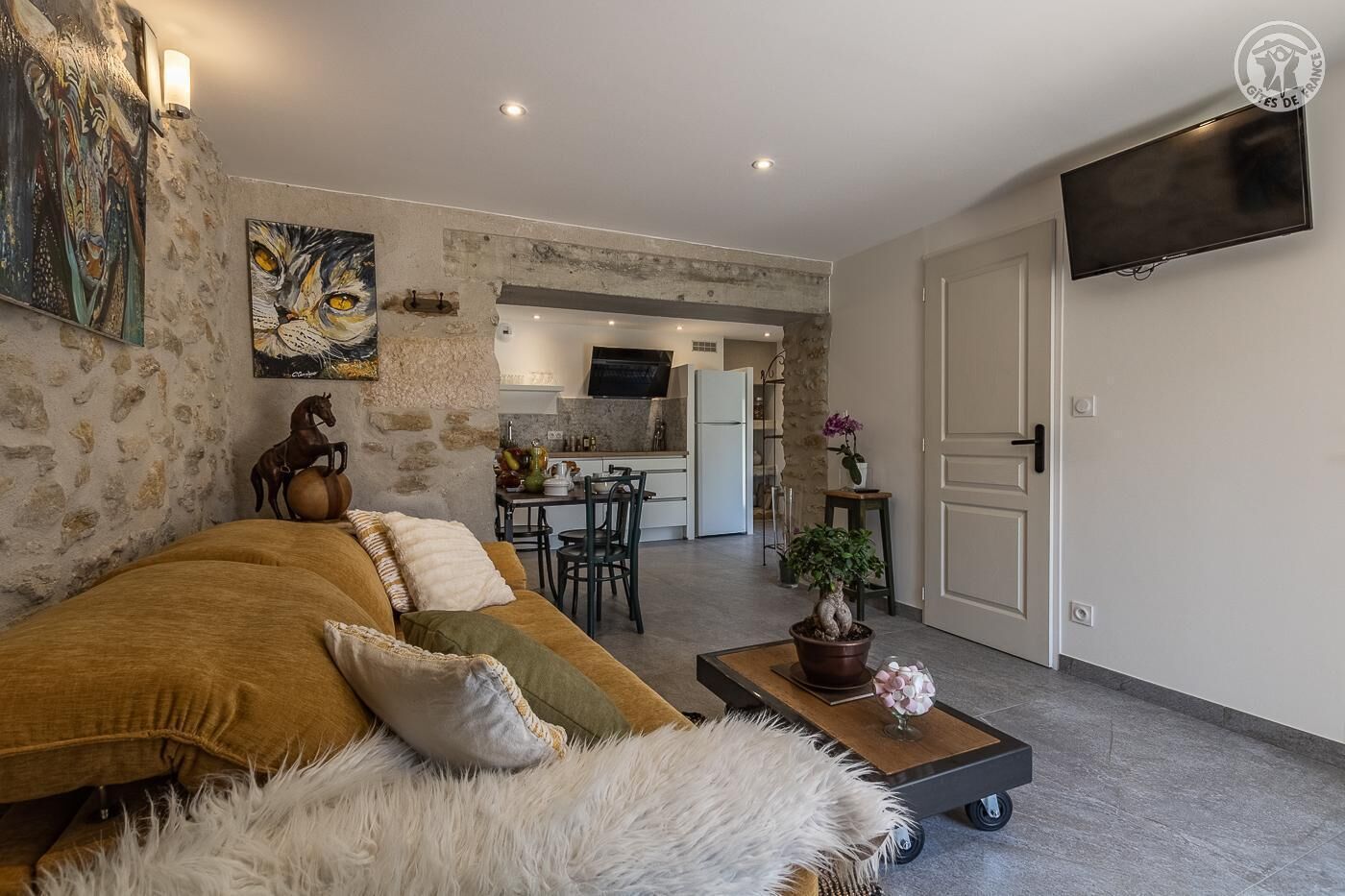 A cozy living room with a sofa, a coffee table, and a TV. There's a kitchen in the background with a dining table and chairs.