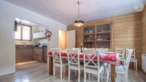 A dining area with a wooden table and chairs, a kitchen in the background, and a wooden wall with shelves. - CHALET PRANA - HAUTELUCE (HAUTELUCE)