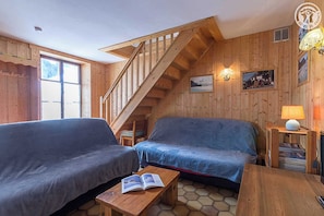 Un interior de madera con dos sofás, una escalera de madera, una mesita con un libro y una lámpara en una mesita auxiliar.