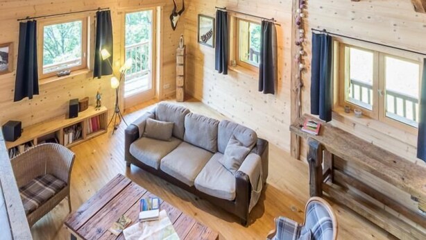 A wooden cabin interior with a high ceiling, a sofa, a table, and a chair.