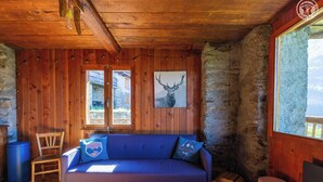 A cozy cabin interior with a wooden ceiling, a blue sofa with matching cushions, a stone wall, and a window with a view of a building outside.