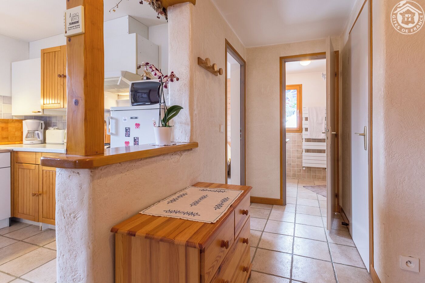 Una cocina con microondas, cafetera y una orquídea en maceta. Hay una encimera de madera con un mantel individual y un pequeño cofre de madera.