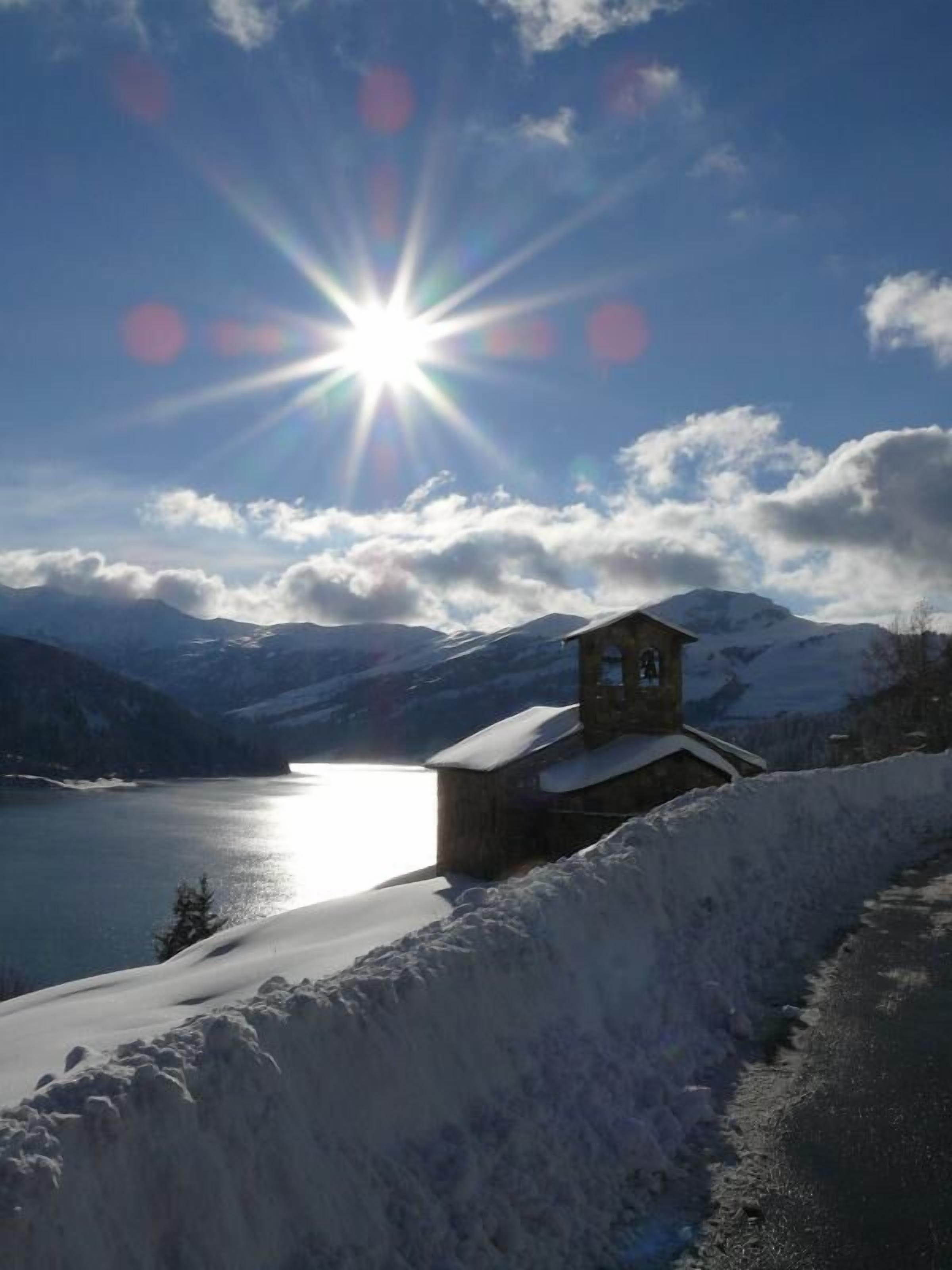 A snow-covered landscape with a small building near a lake, surrounded by mountains.