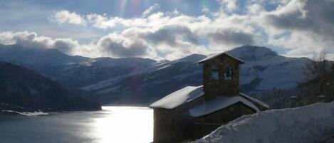 A snow-covered landscape with a small building near a lake, surrounded by mountains.