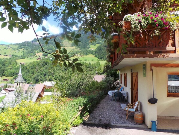 A chalet with a balcony filled with flowers, a gravel path leading to a table and chairs, and a view of a church and green hills in the background.