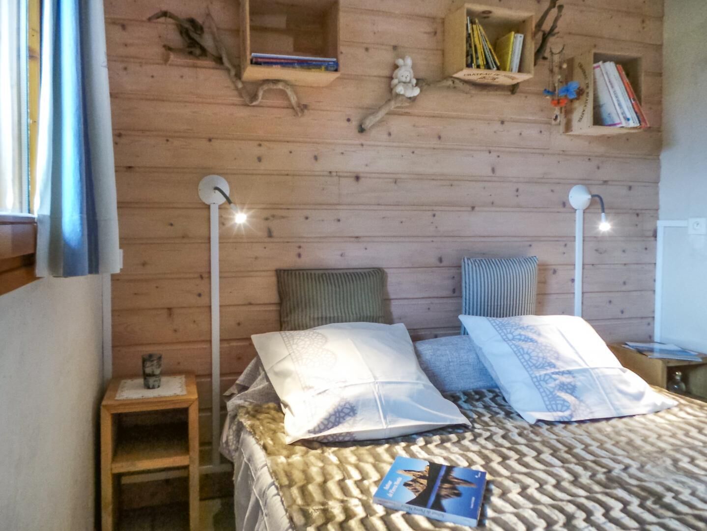 A wooden-paneled bedroom with a bed, two bedside lamps, a small table, and a shelf with books and decorative items.