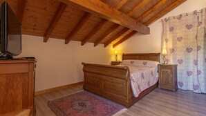 Un dormitorio con techo de madera, una cama con colcha estampada, una mesita de noche con lámpara y un televisor sobre un mueble.