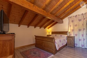 A bedroom with a wooden ceiling, a bed with a patterned bedspread, a nightstand with a lamp, and a television on a cabinet.