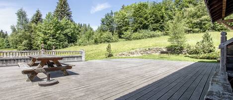 Eine Holzterrasse mit Picknicktisch, umgeben von einem Geländer und Blick auf einen bewaldeten Hügel.