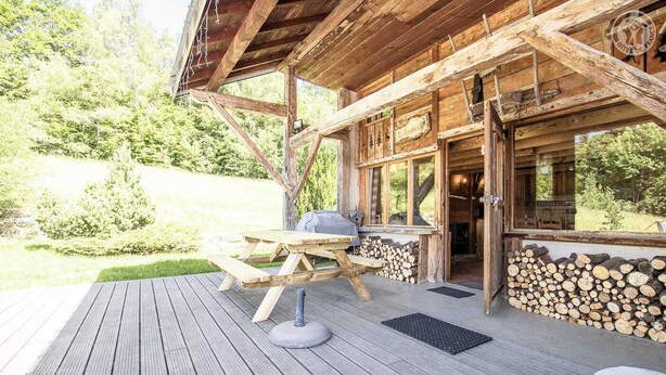 A wooden deck with picnic tables and a barbecue grill, surrounded by a stack of firewood and a view of a forested area.
