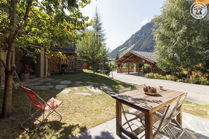 A rustic outdoor seating area with a wooden table and chairs, surrounded by a stone pathway and lush greenery.