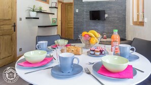 A dining area with a round table set for two, featuring a bowl of fruit, a bottle of juice, and various dishes.
