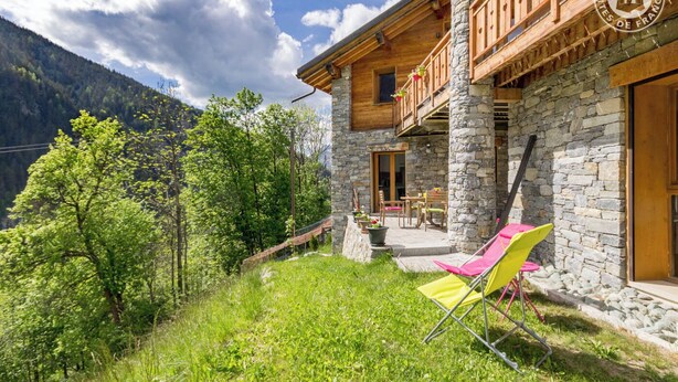 A stone building with a balcony, outdoor seating, and a view of a forested mountain.