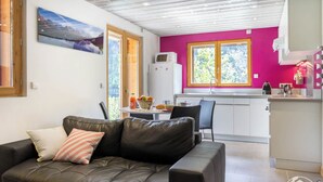 A modern living room with a leather sofa, a dining table with chairs, and a kitchen area with a refrigerator and a window with a pink wall.