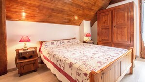 Un dormitorio con techo de madera, una cama con colcha estampada, dos mesitas de noche con lámparas y un armario de madera.