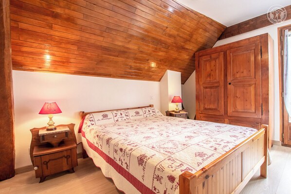 Een slaapkamer met een houten plafond, een bed met een bedsprei met dessin, twee nachtkastjes met lampen en een houten garderobe.