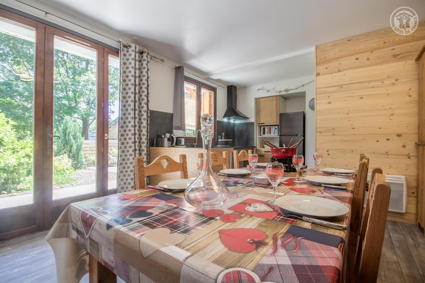 Een eetkamer met een tafel voor vier personen, een keuken op de achtergrond en grote ramen met gordijnen.