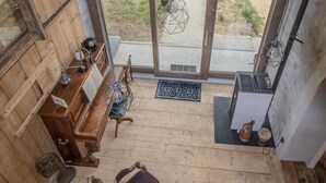 A wooden interior with a piano, two chairs, a small table, and a view of a green landscape through large windows.