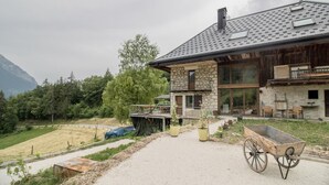 A two-story stone house with a balcony, surrounded by a garden and a view of mountains.