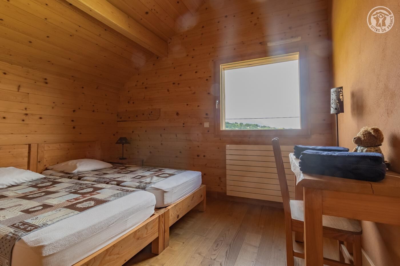 A wooden cabin room with two beds, a table, a chair, a window, and a radiator.