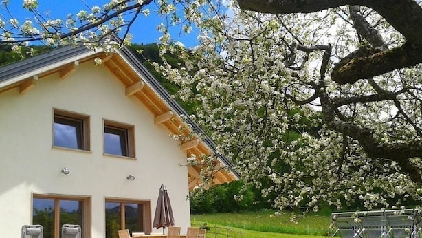 Une maison à deux étages avec un balcon, un coin repas comprenant des chaises et une table, et un jardin avec un arbre en fleurs.