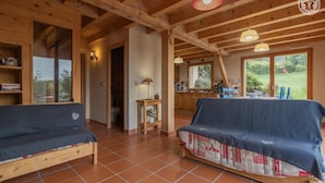 Une pièce chaleureuse avec des poutres apparentes, un canapé, une table et une vue sur un jardin grâce à de grandes fenêtres.