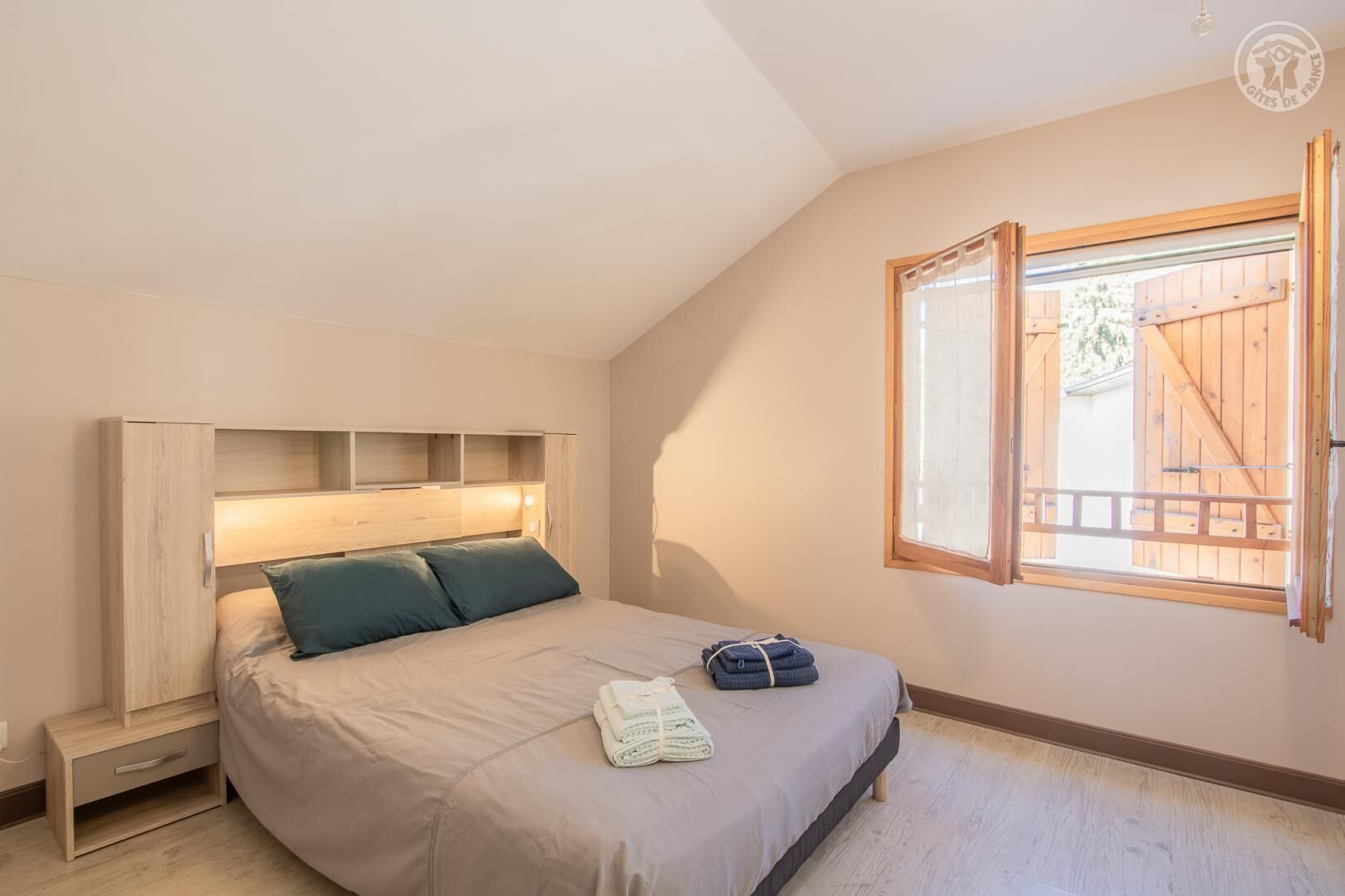 Ein Schlafzimmer mit einem Bett, einem Holz-Kopfteil, einem Fenster mit geöffnetem Rollo und einem kleinen Nachttisch.