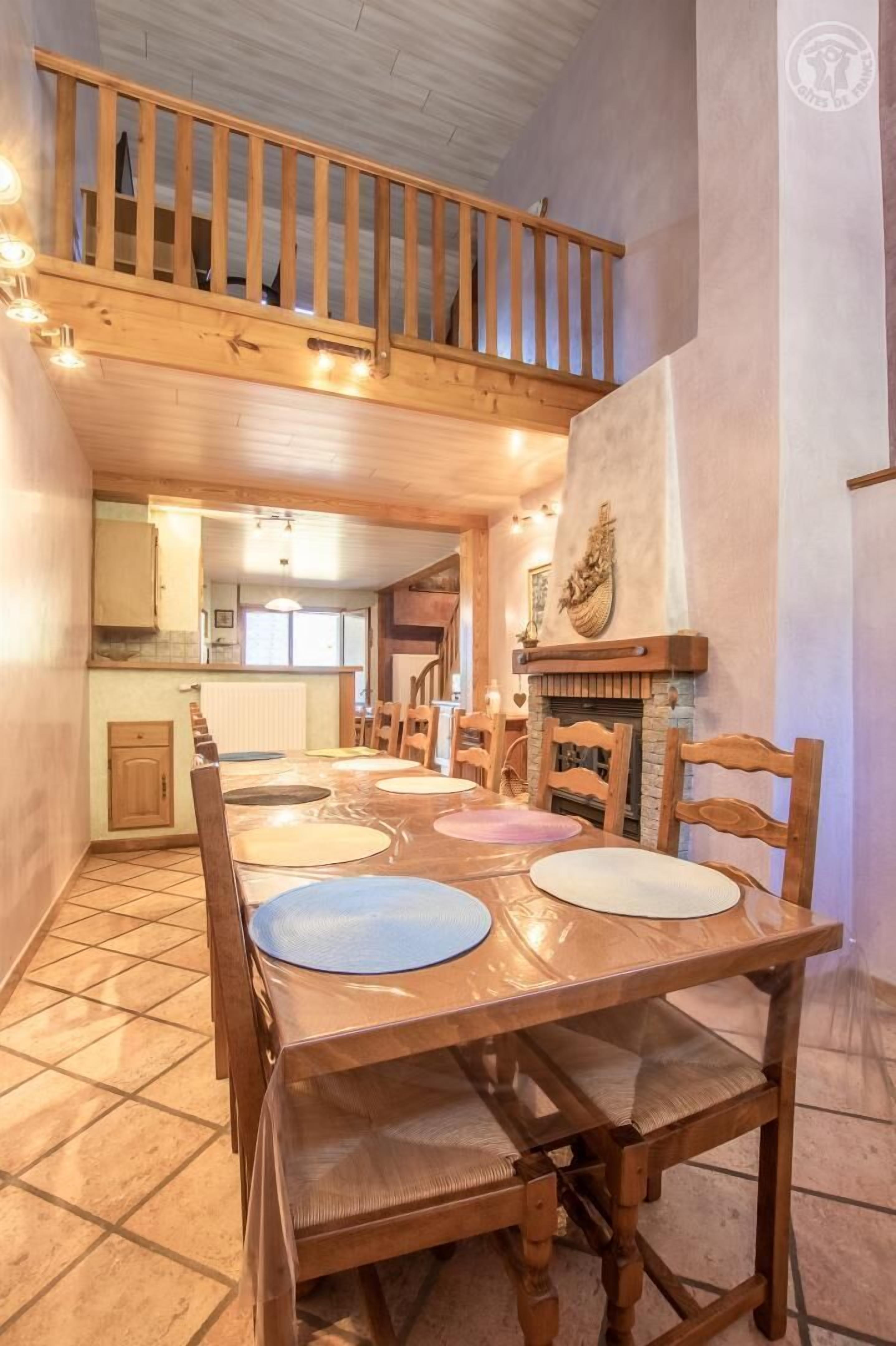 A dining area with a wooden table and chairs, a fireplace, and a balcony with wooden railings.