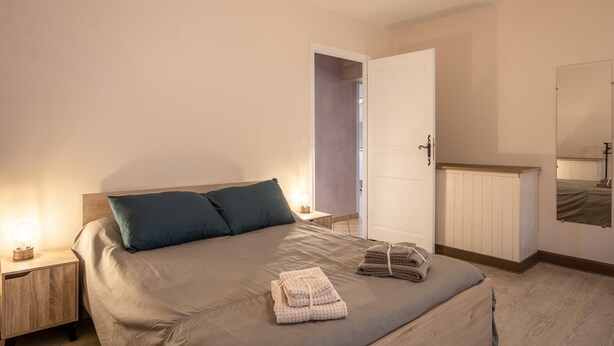 A bedroom with a bed, two pillows, a nightstand, a bedside table, a mirror, and a door leading to another room.