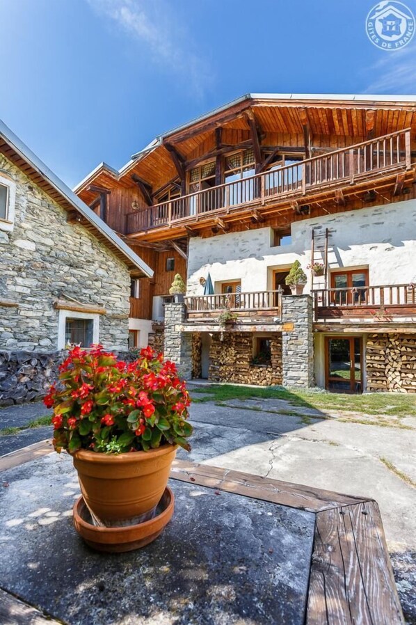 Un bâtiment de style chalet, avec une façade en pierre, des balcons en bois et, au premier plan, un pot de fleurs rouges.