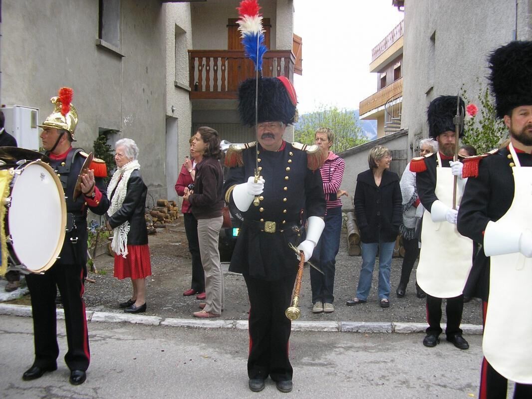 A group of uniformed individuals in ceremonial attire, including a drummer and a person holding a flag, standing on a street with onlookers.