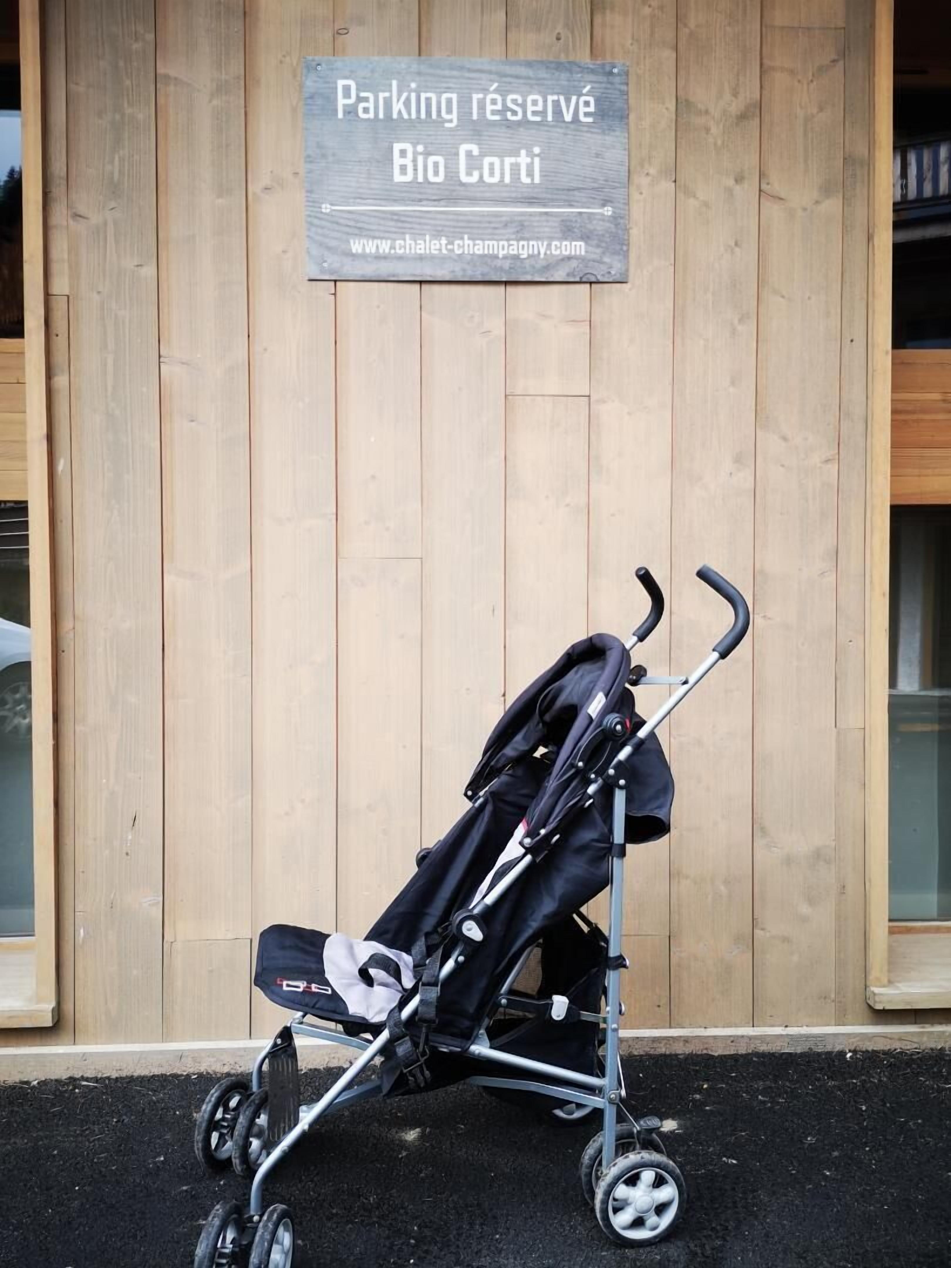 Een kinderwagen geparkeerd voor een houten gebouw met een bord waarop staat 'Parking réservé Bio Corti' en een websiteadres.