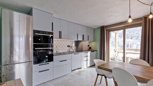 A modern kitchen with white cabinets, a stainless steel Bosch refrigerator, and a dining area with a wooden table and white chairs. - BIO CORTI LODGE - CHAMPAGNY EN VANOISE (CHAMPAGNY EN VANOISE)