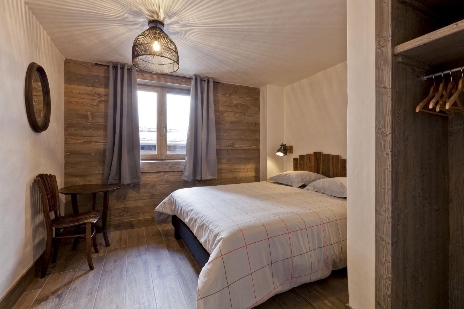 Un dormitorio con suelo de madera, una cama con colcha blanca y roja a cuadros, un espejo redondo, una mesita y una lámpara colgante.