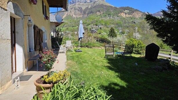 A building with a balcony overlooking a grassy yard, a mountain in the background, and a few outdoor chairs and umbrellas.