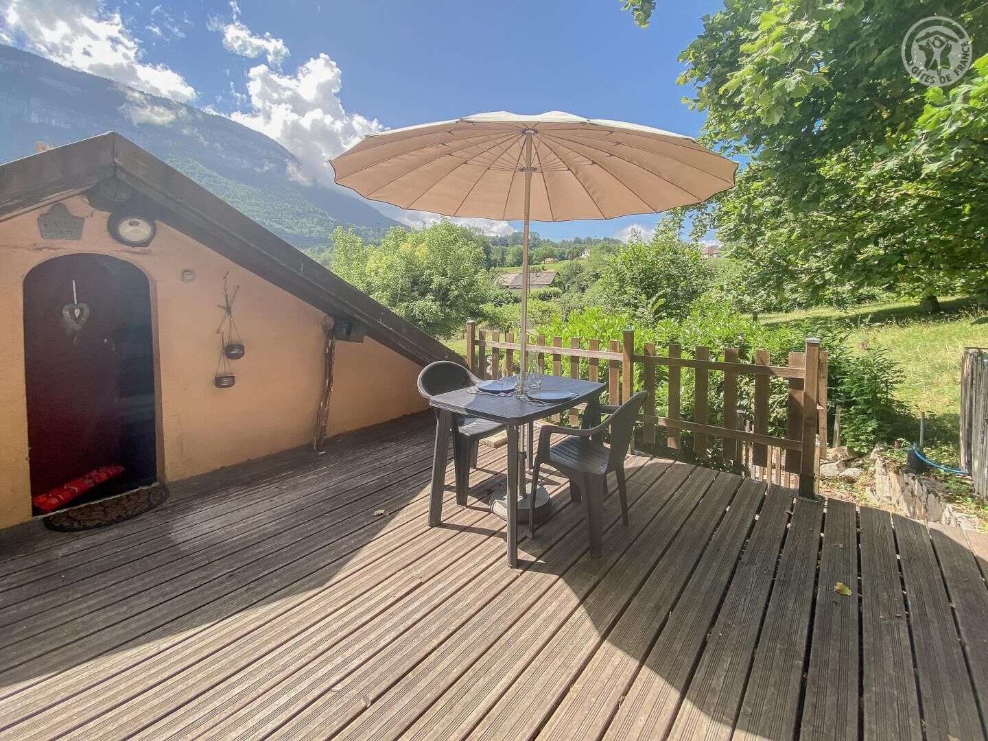A wooden deck with a table and chairs, an umbrella, and a view of greenery and mountains.