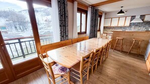 Ein Holzespeisesaal mit sechs Stühlen, eine Küchenzeile mit Holzschränken und große Fenster mit gemusterten Vorhängen.