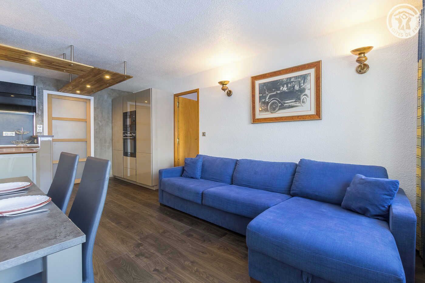 A modern living room with a blue sofa, a dining table with chairs, and a kitchen area.
