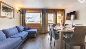 A modern hotel room with a sofa, dining table, chairs, a TV, and a window with a mountain view.