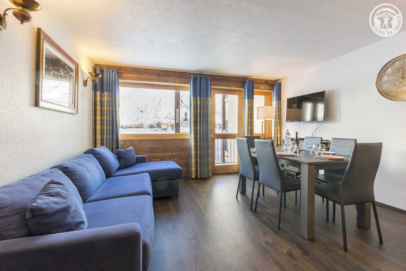 Un salon moderne avec un canapé bleu, une table à manger avec des chaises, un téléviseur fixé au mur et une fenêtre avec des rideaux.