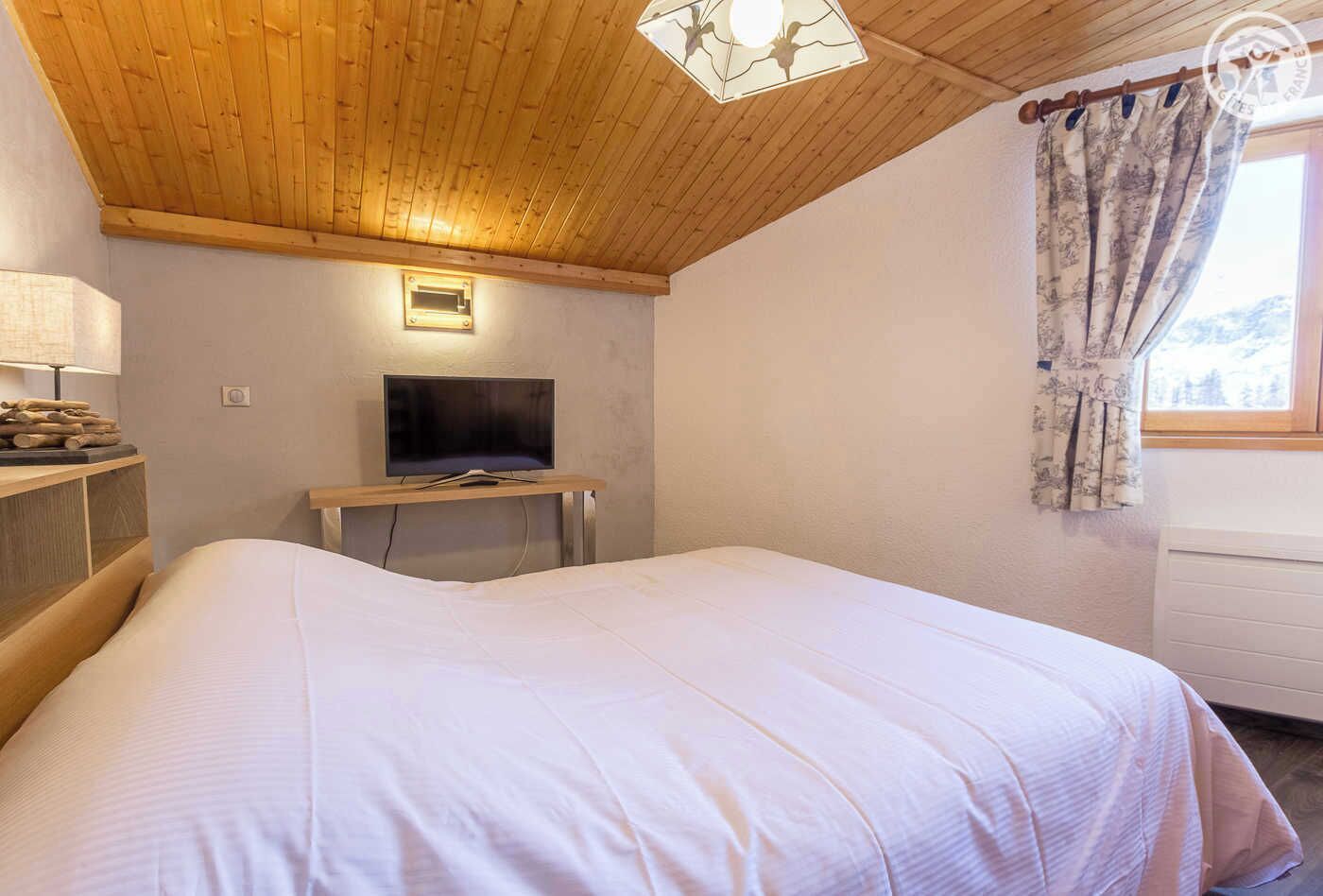 A bedroom with a wooden ceiling, a bed, a TV on a stand, a lamp, and a window with curtains.
